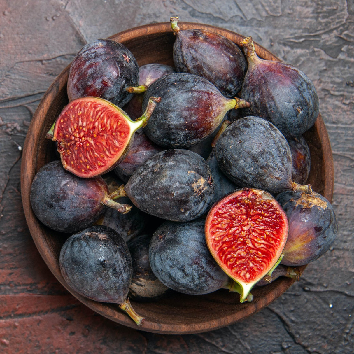 Wooden bowl filled with fresh figs from a Black Mission fig tree, displaying their dark purple skin with a slightly dusty finish. Two figs are cut open, revealing a vibrant red, seed-filled interior that contrasts beautifully with the dark outer skin. The bowl rests on a textured surface, enhancing the rich, rustic look and showcasing the fruit's juicy, fresh appearance.