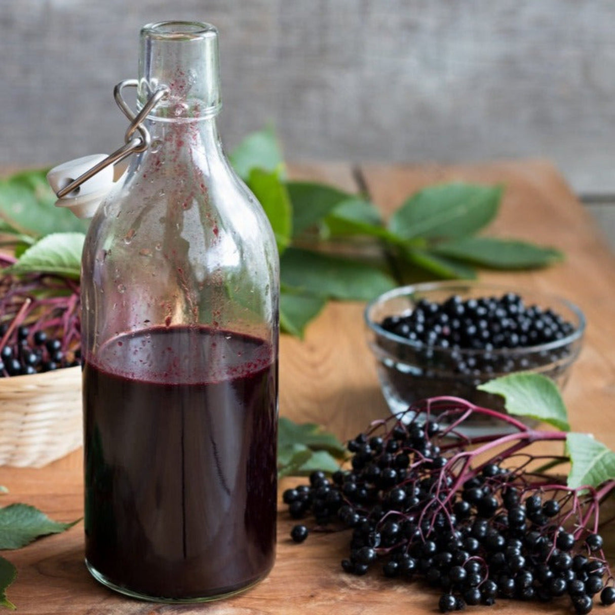 A glass bottle filled with dark elderberry syrup next to fresh elderberries on a wooden surface.