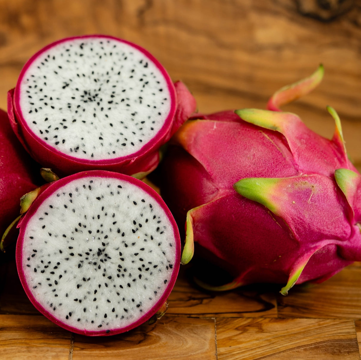 A close-up of ripe dragon fruit, one whole and one sliced in half, showing white flesh with black seeds and vibrant pink skin with green-tipped scales on a wooden surface.