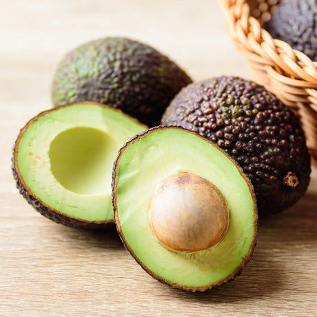 Hass Avocados from the Hass Avocado Tree sitting on a wood countertop. One of the avocados is sliced open revealing the creamy green flesh and light brown seed.