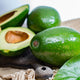 Close-up of whole and halved cold hardy green avocados with smooth, shiny skins and creamy flesh on a wooden surface.